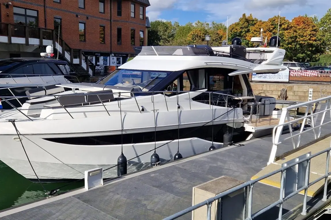 Luxury white motor yacht docked at a marina, with nearby buildings and autumn trees in the background.